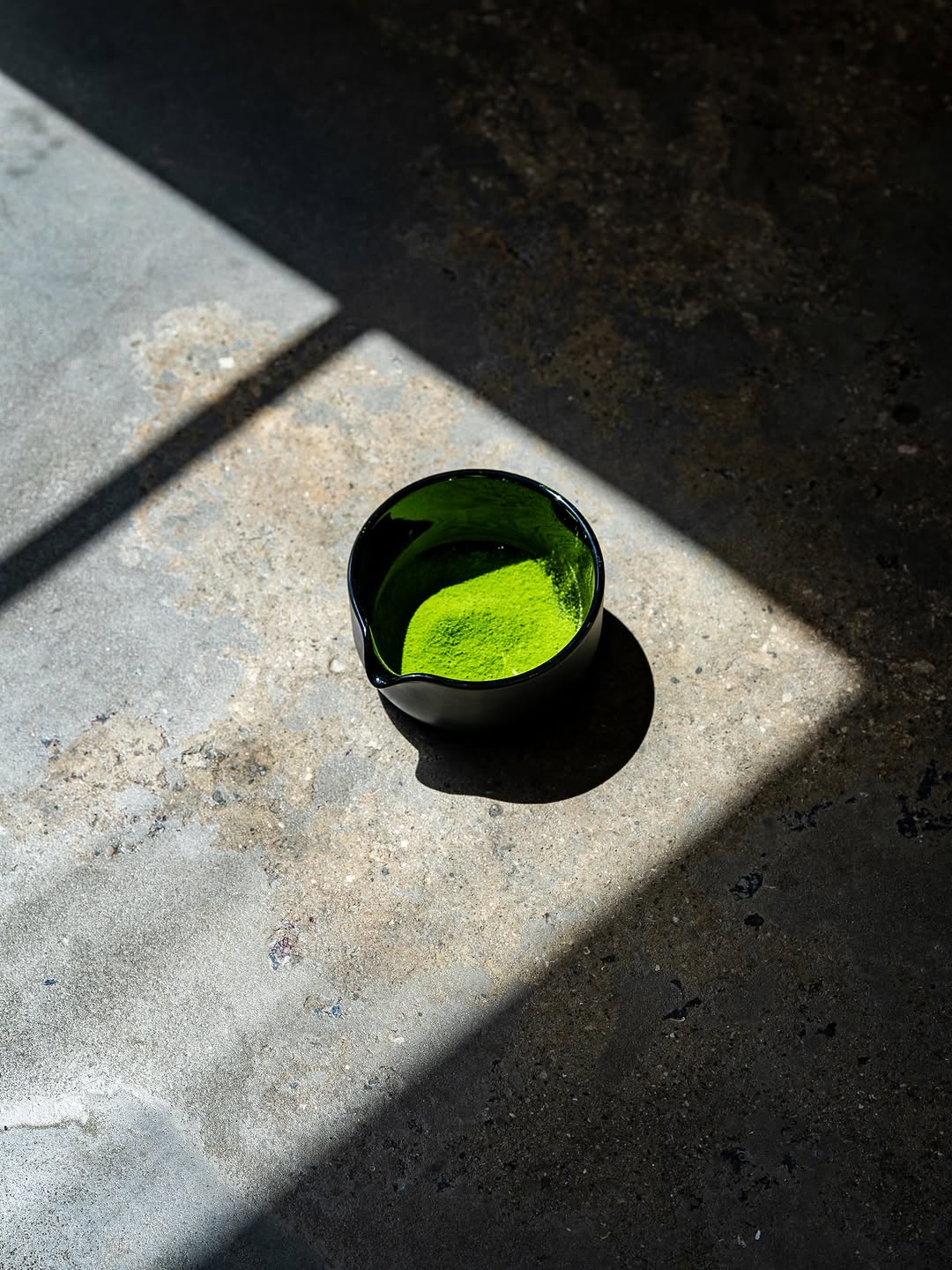 Bright green matcha in a black bowl on concrete, lit by hard sunlight